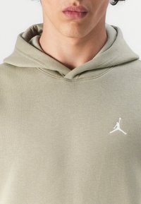 Alyvuogių spalvos megztinis su kapišonu, pagamintas iš minkštos medžiagos, su priekinėmis kengūros kišenėmis ir balta Jumpman logotipo etiketė kairėje krūtinės srityje.
