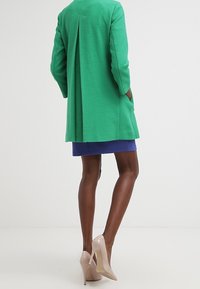 Zelený texturovaný kabát s zadním sklady, kombinovaný s modrou sukní a béžovými vysokými podpatky, což vytváří přiléhavý siluetu.