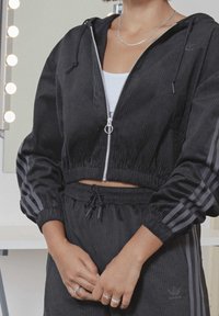 Händerna ihopknutna framför kroppen i en svart Adidas corduroy träningsdräkt med kort zip-jacka och muddbyxor, vit linne syns under.