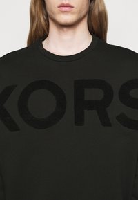 Felpa nera con scollo a costine, caratterizzata da un grande lettering testurizzato "KORS" in un materiale nero a contrasto. Vestibilità casual, tessuto morbido.
