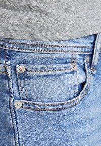 Närbild av ljusblå denimjeansficka med kopparnitar och en del av en grå skjorta ovanför midjebandet.