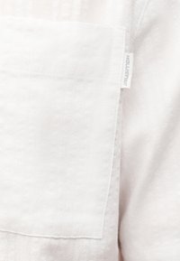 Κοντινή λήψη μιας λευκής υφασμάτινης τσέπης με μια μικρή ετικέτα της μάρκας Hollister ραμμένη στη πλευρική ραφή.