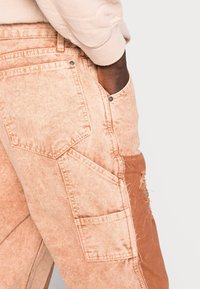 Persona de piel oscura vistiendo jeans de lavado ácido rosa claro con múltiples bolsillos y un detalle rasgado, con la mano dentro del bolsillo delantero.