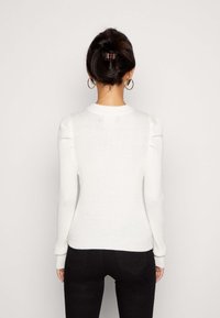 Pull blanc côtelé à manches longues avec un col rond. Présente des détails subtils aux épaules et des poignets ajustés. Porté avec un pantalon noir.