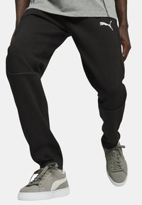 Sorte sweatpants med hvid PUMA-logo, med en tætsiddende design, kontrasterende teksturerede paneler og elastiske manchetter, parret med grå sneakers.