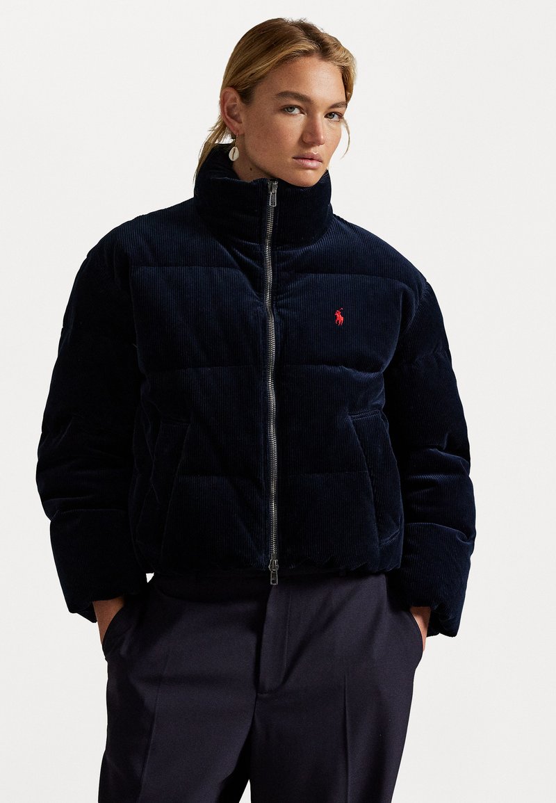 Marineblauwe corduroy pufferjack met hoge kraag, ritssluiting en rood logo op de borst. Heeft gevoerde delen en zijzakken.