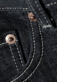Denimstoff i mørkeblått med synlig hvit søm, detaljer i kobbernitter og en liten lommeåpning. Teksturert med en tett vev.