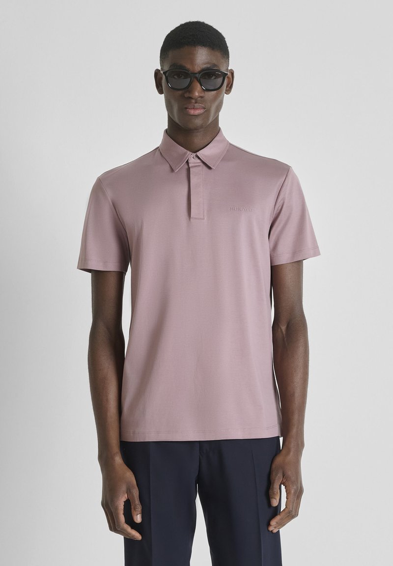 Hombre con gafas de sol oscuras, polo de manga corta color malva y pantalones oscuros, de pie frente a un fondo claro y liso.