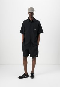 Sort kortærmet skjortebluse med knapper og matchende shorts, begge med en lomme med "GAP" logoen. Sættet er kombineret med sorte sneakers og en grå kasket.