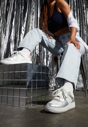 Baskets blanches et argentées avec des surfaces texturées, des accents roses et des lacets. Le modèle porte un jean bleu clair et des chaussettes blanches, assis sur un cube réfléchissant.