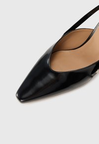 Zwarte patent leren slingback schoen met een puntige neus, voorzien van een gladde textuur en open zijkanten, die een beige binnenzool onthullen.