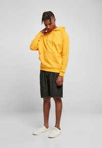 Sudadera amarilla con un bolsillo frontal, combinada con pantalones cortos de mezclilla oscura y zapatillas blancas. El modelo está de pie frente a un fondo neutro, mirando hacia abajo.