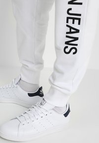 Białe spodnie dresowe z czarnym napisem „STAN JEANS” na lewej nogawce, do tego białe buty adidas Stan Smith i szare skarpetki.