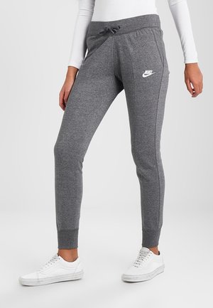 Persoon draagt grijze Nike joggingbroek met trekkoord in de taille en witte sneakers, staand tegen een effen achtergrond.