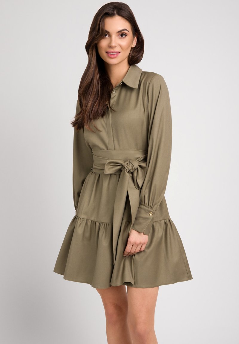 Oliwkowa sukienka z długim rękawem, kołnierzykiem zapinanym na guziki, wiązaniem w talii i falbanistym dołem. Gładka tkanina i delikatny połysk podkreślają design.