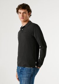 Černá polokošile s dlouhým rukávem vyrobená z texturované látky, s tříknoflíkovým zapínáním a decentním logem na hrudi, v kombinaci s modrými džíny.