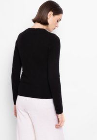 Maglione nero lavorato a maglia con maniche lunghe, scollatura rotonda e una silhouette aderente. La texture appare morbida e liscia su uno sfondo chiaro.