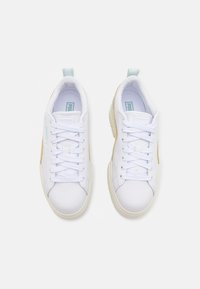 Bílé tenisky s hladkým koženým svrškem, texturovanými tkaničkami, světlými tanovými akcenty po stranách a bílou gumovou podrážkou. Diskrétní značení na jazyku.