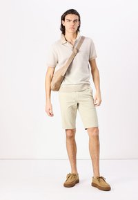 Beige Poloshirt mit strukturiertem Muster, helle khakifarbene Shorts und braune Wildlederschuhe. Das Modell trägt eine Umhängetasche. Schlichtes, lässiges Design.