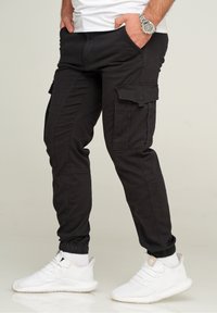 Schwarze Cargo-Hosen mit elastischen Bündchen, ausgestattet mit Seitentaschen und einem eleganten Design. Getragen mit weißen Sportschuhen und einem weißen T-Shirt.