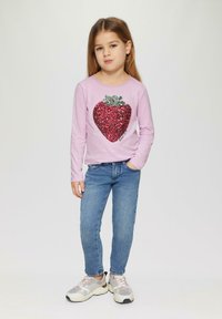 Roze overhemd met lange mouwen met een groot rode pailletten hartontwerp. Gecombineerd met blauwe jeans en lichte sneakers. Eenvoudige, casual outfit.