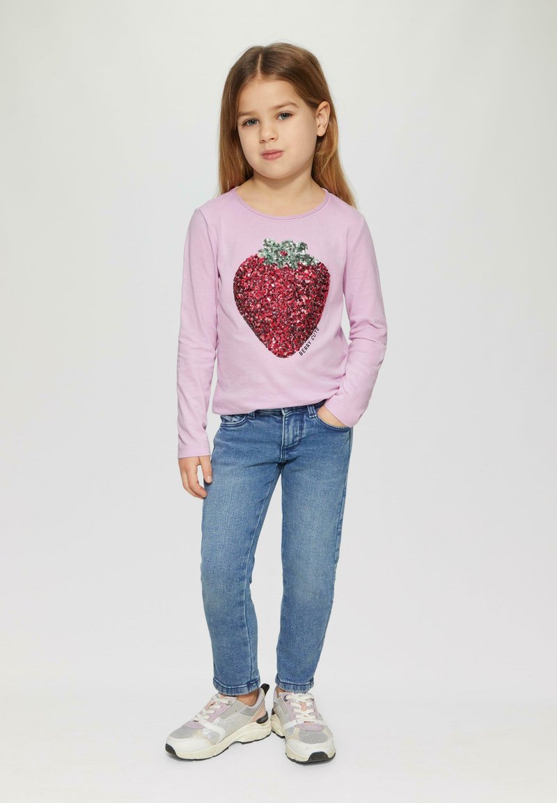 Roze overhemd met lange mouwen met een groot rode pailletten hartontwerp. Gecombineerd met blauwe jeans en lichte sneakers. Eenvoudige, casual outfit.
