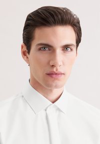 Młody mężczyzna z starannie ułożonym ciemnym włosem, ubrany w białą zapiętą koszulę, patrzący prosto przed siebie z neutralnym wyrazem twarzy.