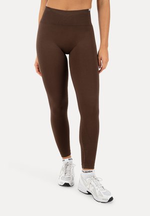 Kobieta ubrana w brązowe legginsy z wysokim stanem, białe skarpetki z logo i białe sportowe buty, stoi na tle jednolitego tła.