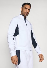 Fehér, cipzáros sportdzseki tengerészkék oldalpanellel, magas gallérral és struktúrált felülettel. Bal oldalon cipzáras zseb található.