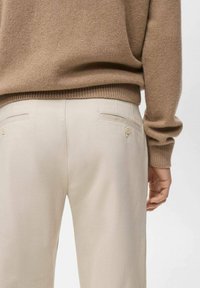 Pantalones beige a medida con dos bolsillos traseros con cierre de botón. Combinados con un suéter marrón de punto. Textura suave y diseño ajustado.