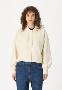 Krämliknande kort jacka med texturerad tyg, som har två framfickor och en ståkrage. Bärs med blå denimjeans.