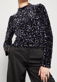 Top nero con paillettes e maniche lunghe, caratterizzato da pailettes viola e argento in un motivo denso. Collo alto e design aderente. Indossato con pantaloni neri.