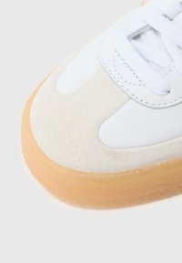 Baskets blanches avec une tige en cuir lisse et des accents en daim. Semelle en caoutchouc couleur beige clair et lacets blancs. Design arrondi et minimaliste.