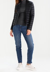 Svart vadderad jacka med figursydd design, dragkedja och guldfärgad metall. Bärs över en mörk polotröja, kombinerad med blå jeans och vita sneakers.