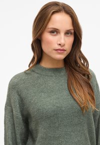 Zielony prążkowany sweter z wysokim kołnierzem, z długimi rękawami i miękkim, teksturowanym wyglądem. Gładkie wykończenie i luźny krój.