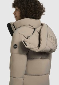 Beige puffjakke med høj krave, horisontalt quiltet tekstur, en mærkebeskyttelse, og lynlåslommer på siden. Holdbart materiale og elegant design.