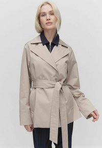 Beżowy trench coat z paskiem, z szerokimi klapami, luźnymi rękawami i dwiema bocznymi kieszeniami, noszony na ciemnoniebieskiej koszuli z zamkiem. Gładki, lekki materiał.