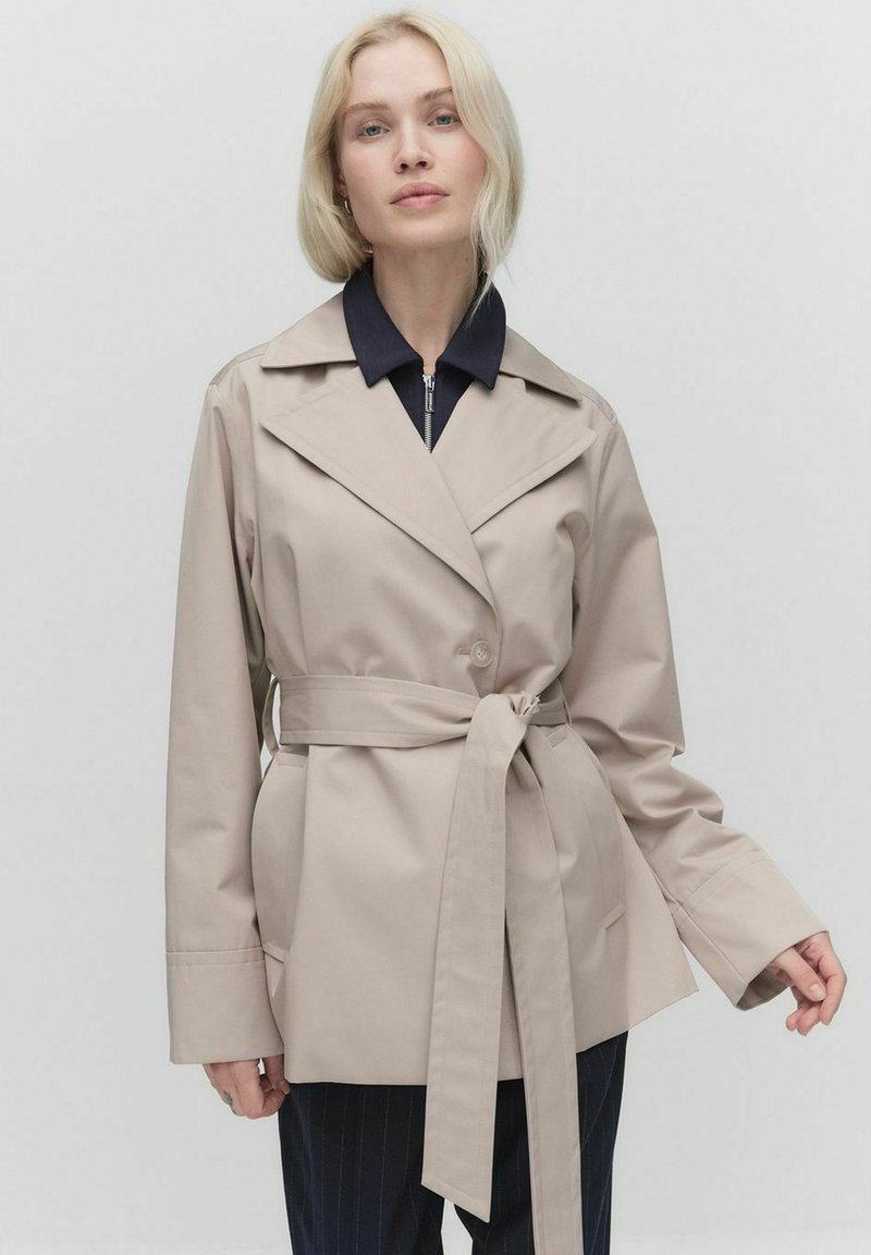 Beżowy trench coat z paskiem, z szerokimi klapami, luźnymi rękawami i dwiema bocznymi kieszeniami, noszony na ciemnoniebieskiej koszuli z zamkiem. Gładki, lekki materiał.