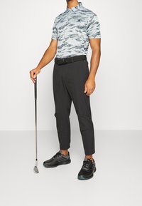 Golfové oblečení obsahující světle modrou patterned polo košili, černé zúžené kalhoty a černé golfové boty s modrými akcenty, držící golfovou hůl.