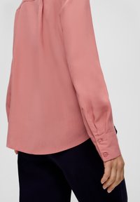 Helle rosa Bluse mit langen Ärmeln, entspannter Passform, Knopfmanschetten und leichtem Glanz; kombiniert mit dunkelblauen Hosen.