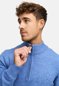 Lyseblå, struktureret zip-up sweater med høj krave og ribbede manchetter. Lynlåsen er delvist åben, hvilket afslører en stolpe.