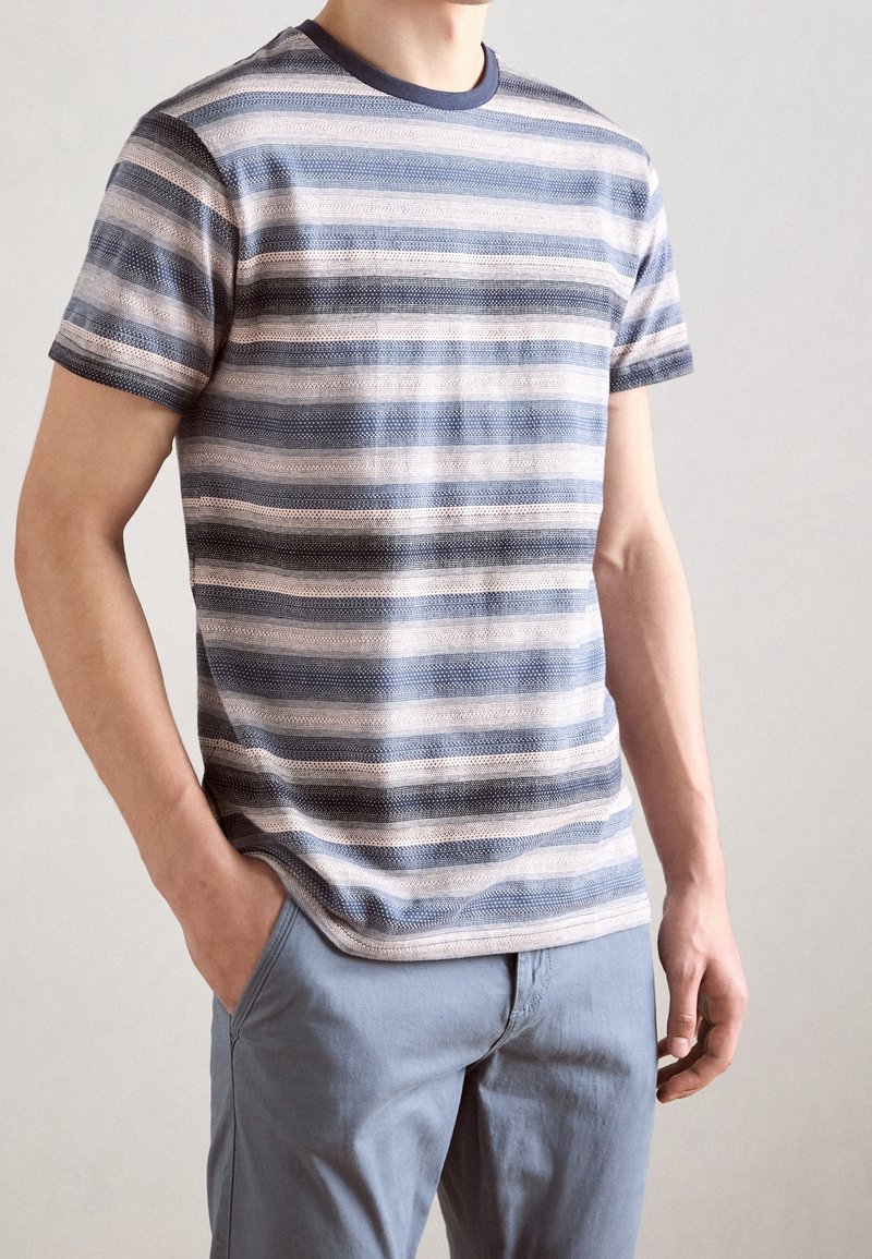 Homme portant une chemise à manches courtes rayée horizontalement bleu et beige avec col bleu, et un pantalon bleu clair avec la main gauche dans la poche.