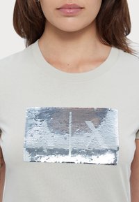 Světle šedé bavlněné tričko s obdélníkovým stříbrným flitrovým vzorem na hrudi, který poskytuje texturovaný, reflexní povrch.