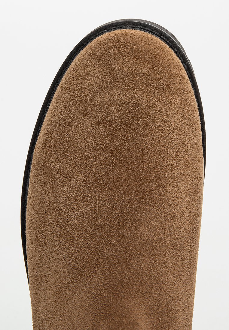 Zbliżenie brązowego zamszowego buta z czarną gumową podeszwą, ukazujące teksturę i detale materiału na białym tle.