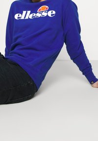 Sweat-shirt bleu avec un grand logo blanc "ellesse" et un accent coloré. Encolure arrondie et manches longues, assorti à un jean foncé.