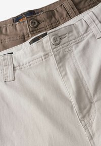 Dos pares de pantalones: uno gris claro y otro marrón. Ambos tienen una textura suave, cierres de botón y detalles de costura visibles.