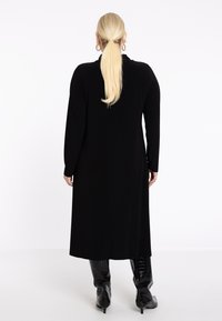 Ein langes schwarzes Kleid aus glattem Stoff, mit langen Ärmeln und geradem Schnitt, kombiniert mit glänzenden knielangen schwarzen Stiefeln.