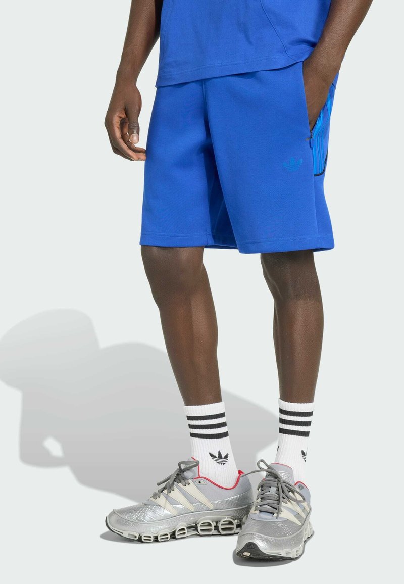 Homme portant un short bleu Adidas, des chaussettes blanches Adidas mi-mollet avec des rayures noires et des chaussures de running argentées Adidas sur un fond clair.