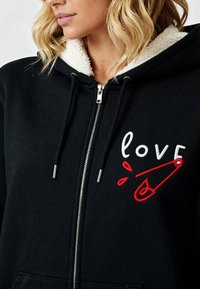 Haut à capuche noir zippé avec un col en sherpa blanc. Présente un texte brodé "love" et un motif de pinces à sécurité rouge.