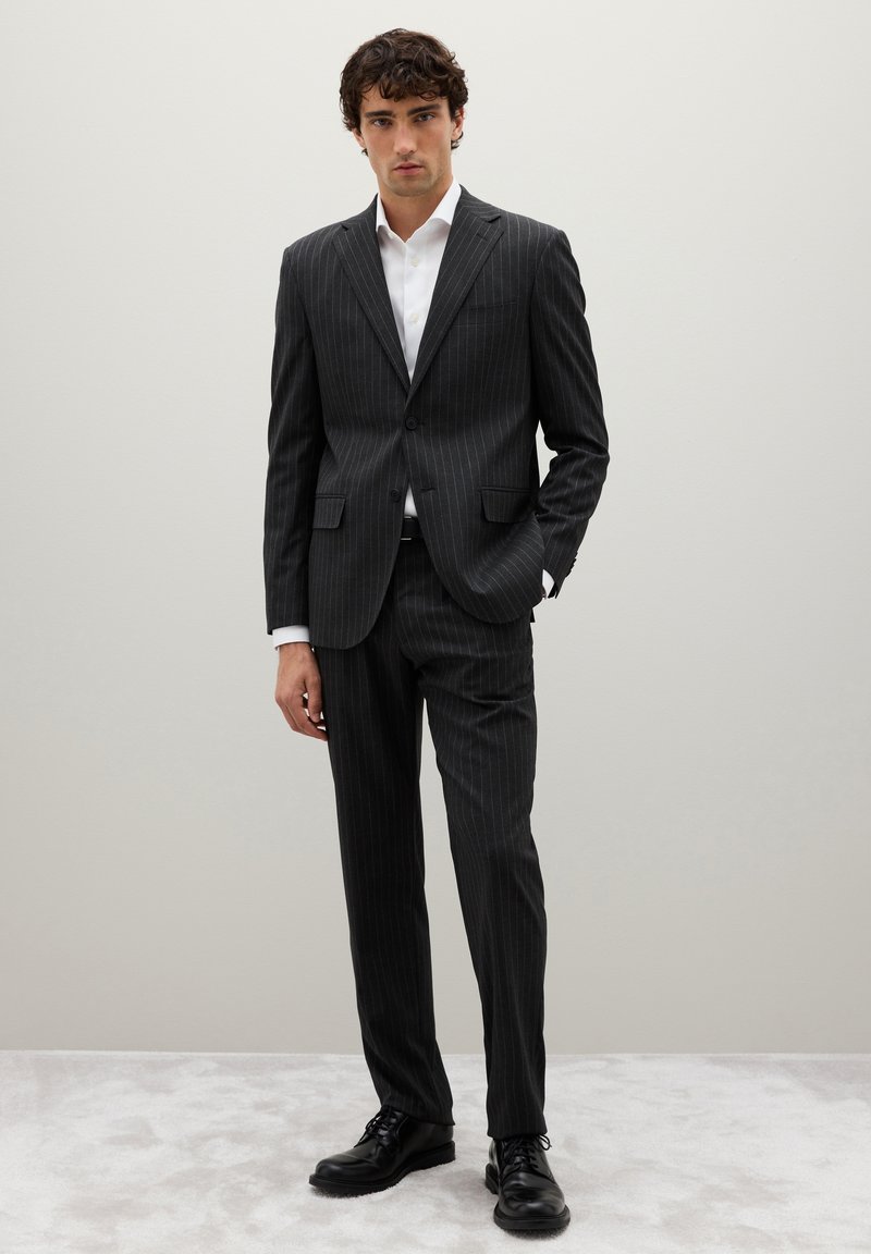 Traje de rayas en carbón con un blazer ajustado y pantalones a juego, combinado con una camisa blanca y zapatos negros de cordones. Textura de tela suave.